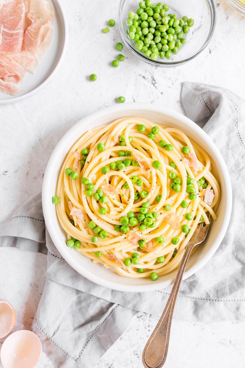 Light Pasta Carbonara with peas and prosciutto on a white plate with eggs in the background