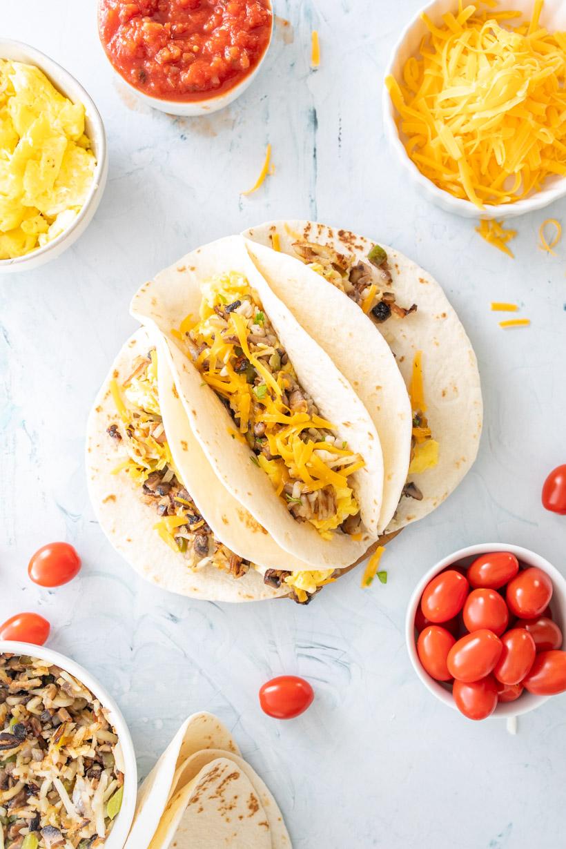 3 tacos filled with eggs and cheese on a plate with ingredients surrounding them