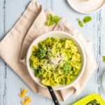 bowtie pasta with pesto in a white bowl with ingredients on table