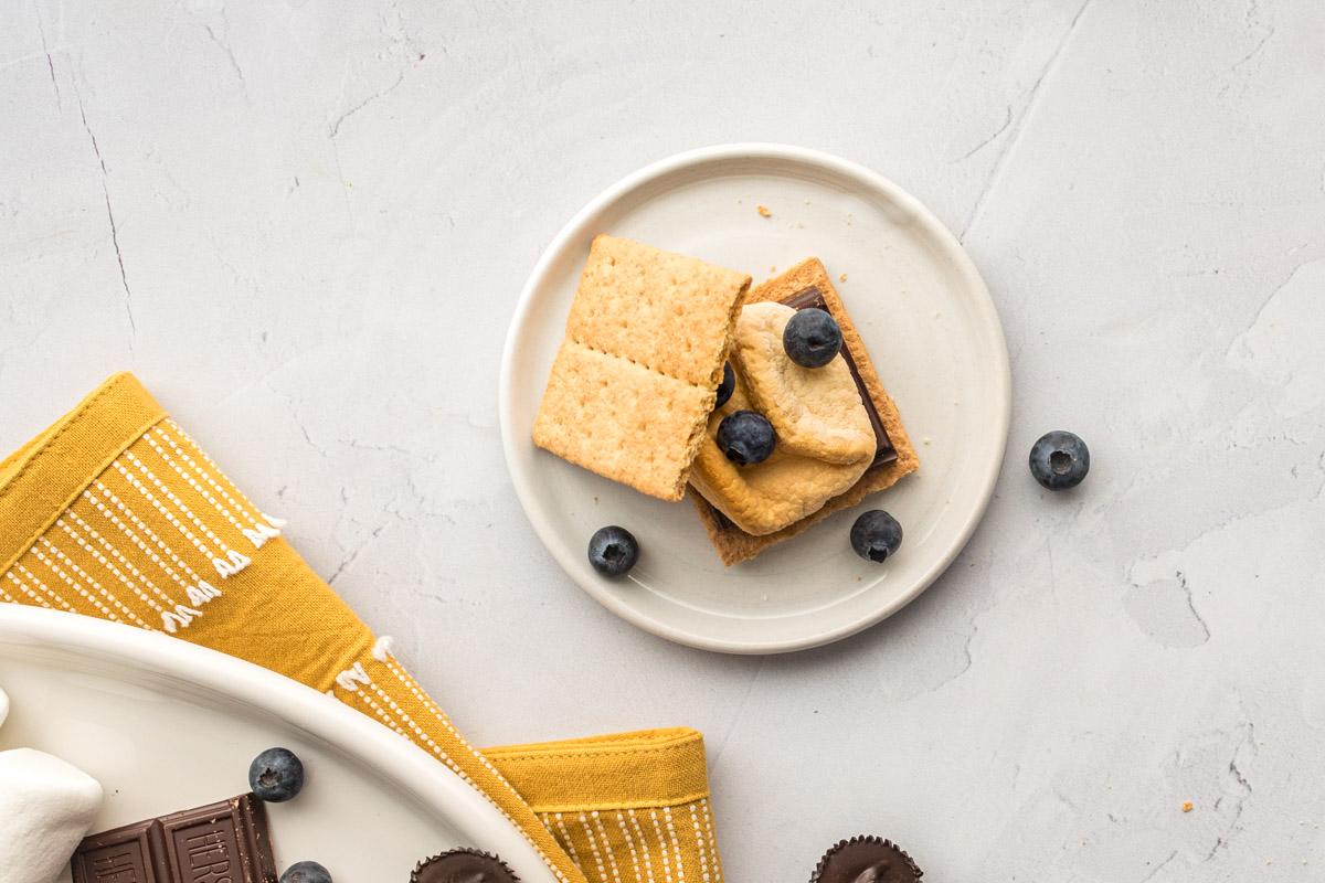 smore with blueberries on a plate with ingredients and yellow towel next to it