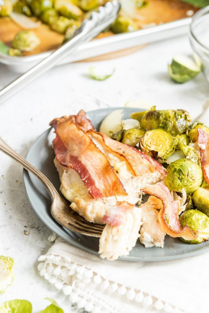a forkful of chicken and bacon with a chicken breast with bacon on top next to brussels sprouts on a plate with a sheetpan in the background