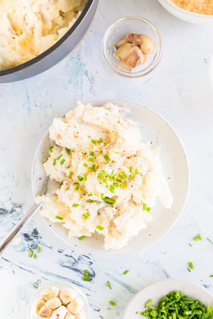 Cheesy garlic mashed potatoes with chives and pepper in a white bowl with ingredients surrounding it