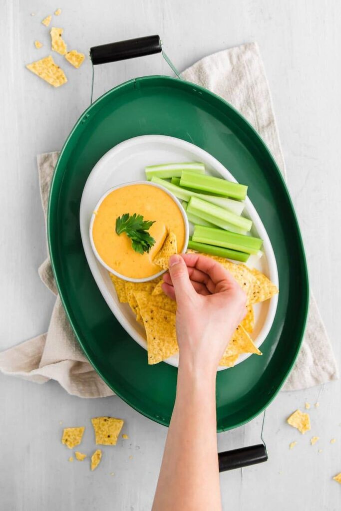 dipping a chip into easy queso dip with chips and celery on a table