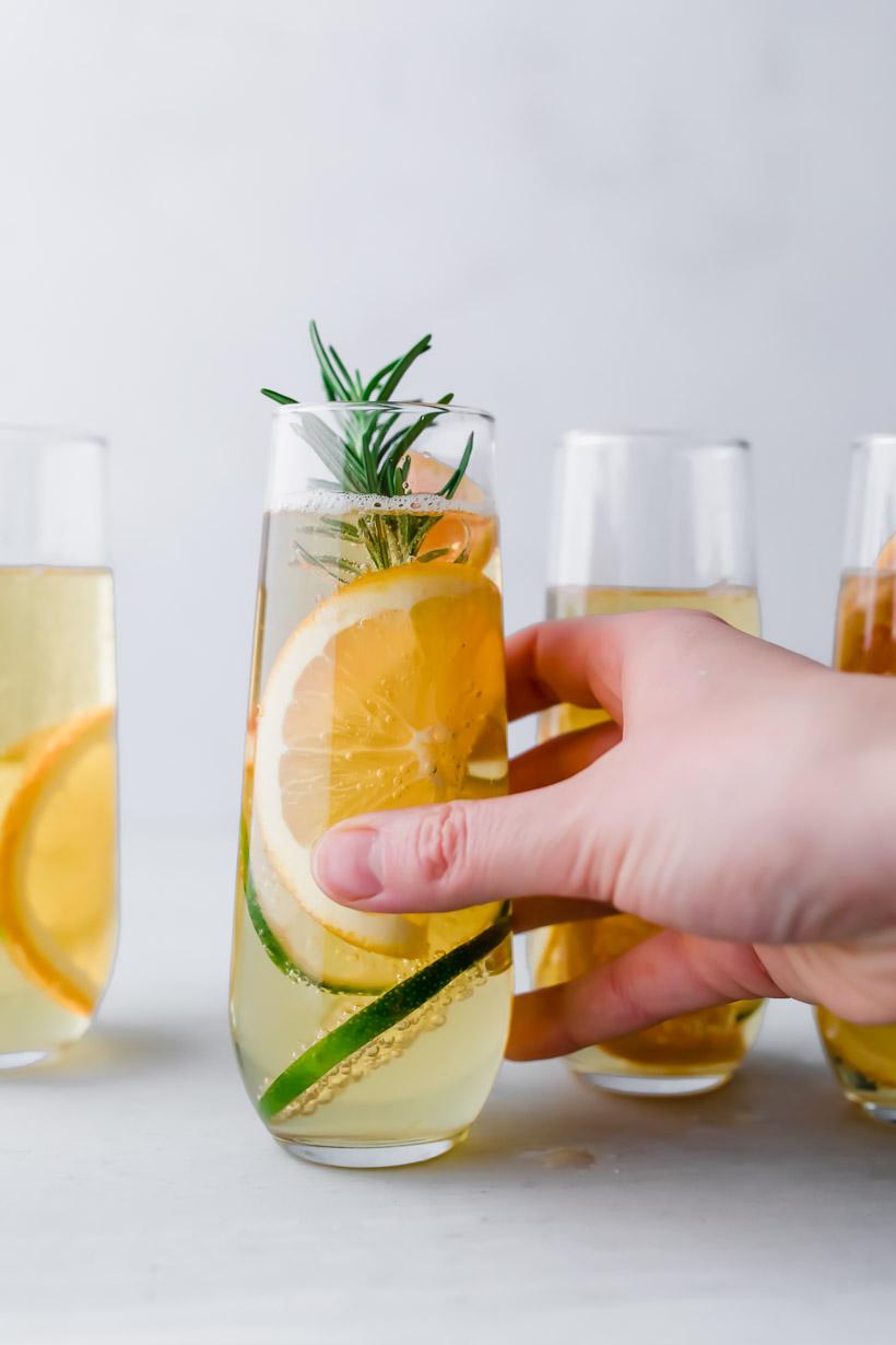 holding an apple drink with sliced citrus and rosemary in a stemless champagne glass