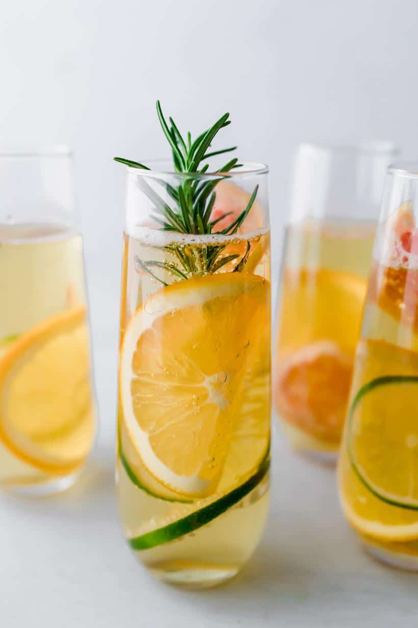 four apple mocktails in stemless champagne glasses with thin slices of citrus and rosemary in each glass