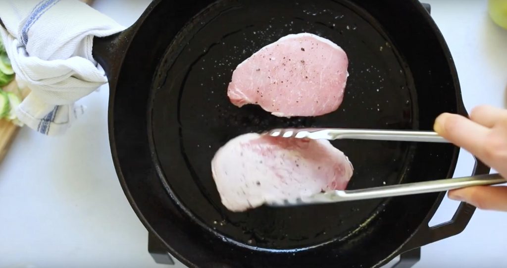 tongs holding one pork chop next to a second pork chop in a cast iron skillet