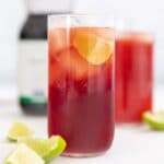 a tall glass of pomegranate juice with lime wedges in it and around it