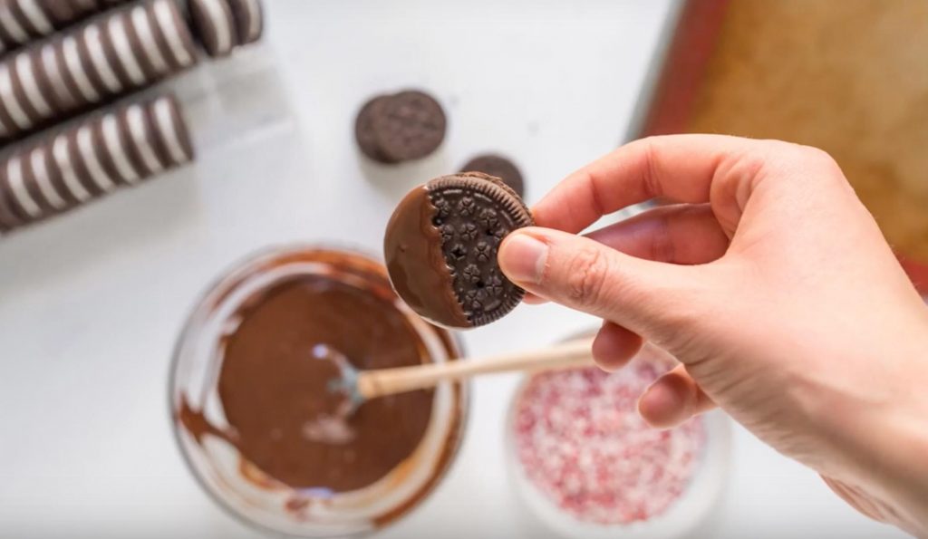 hand holding a chocolate dipped oreo with chocolate, crushed peppermint and oreos below on table