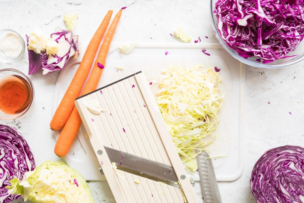 mandolin and shredded cabbage on a cutting board next to coleslaw ingredients