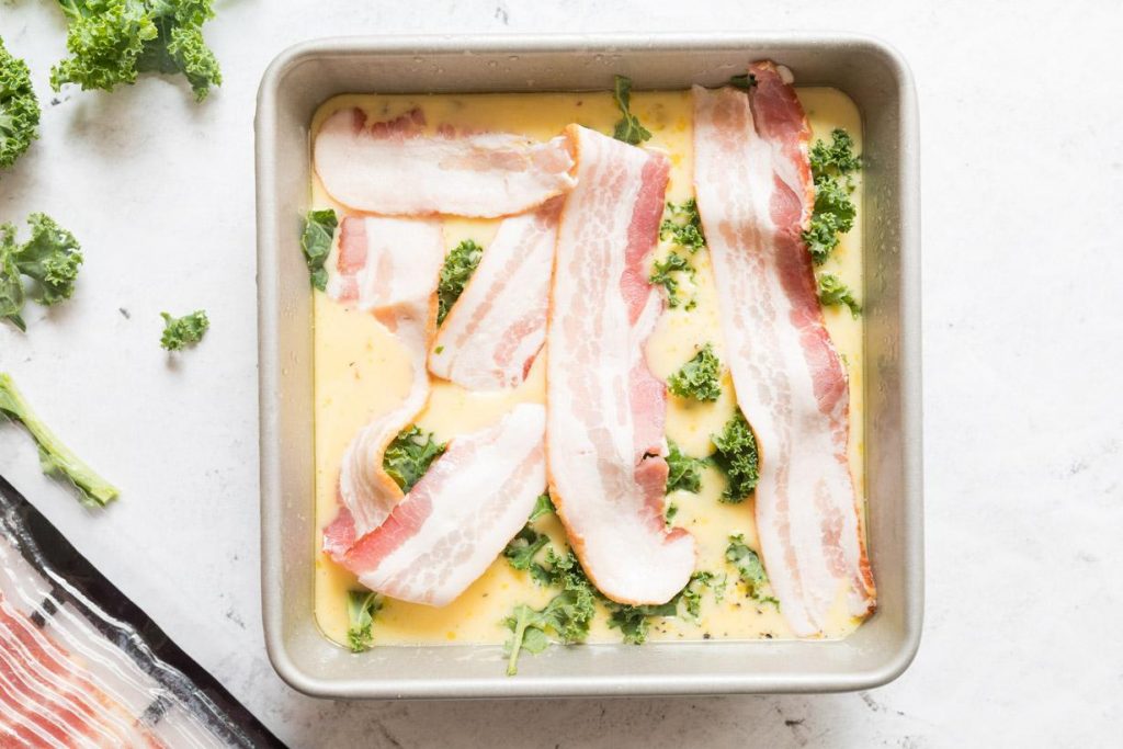 uncooked eggs, kale, and bacon slices in a square baking pan