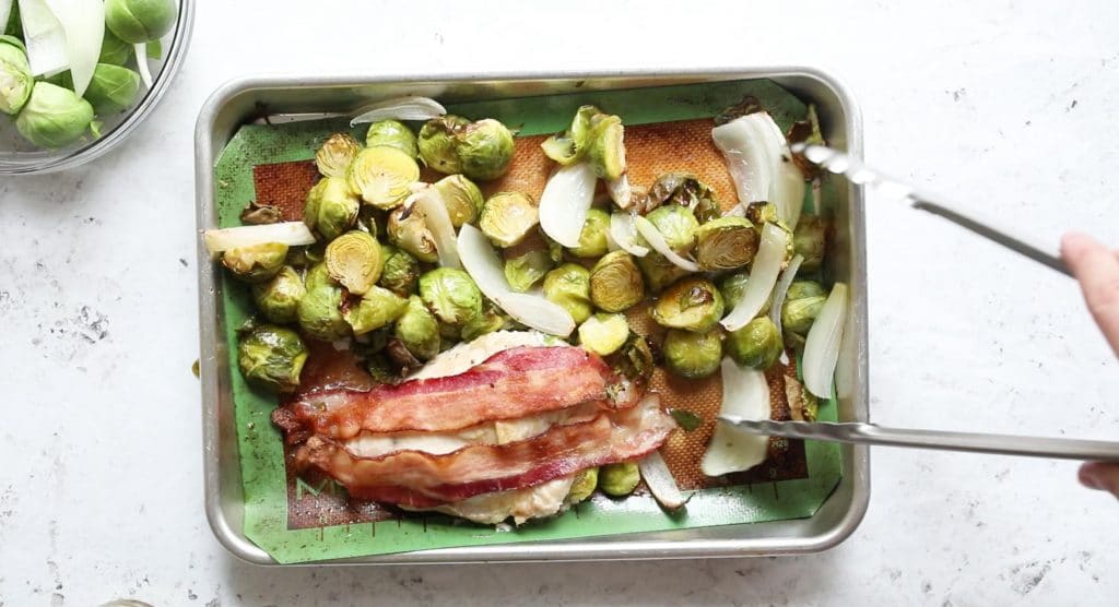cooked bacon on top of chicken with brussels sprouts and onions on a cookie sheet