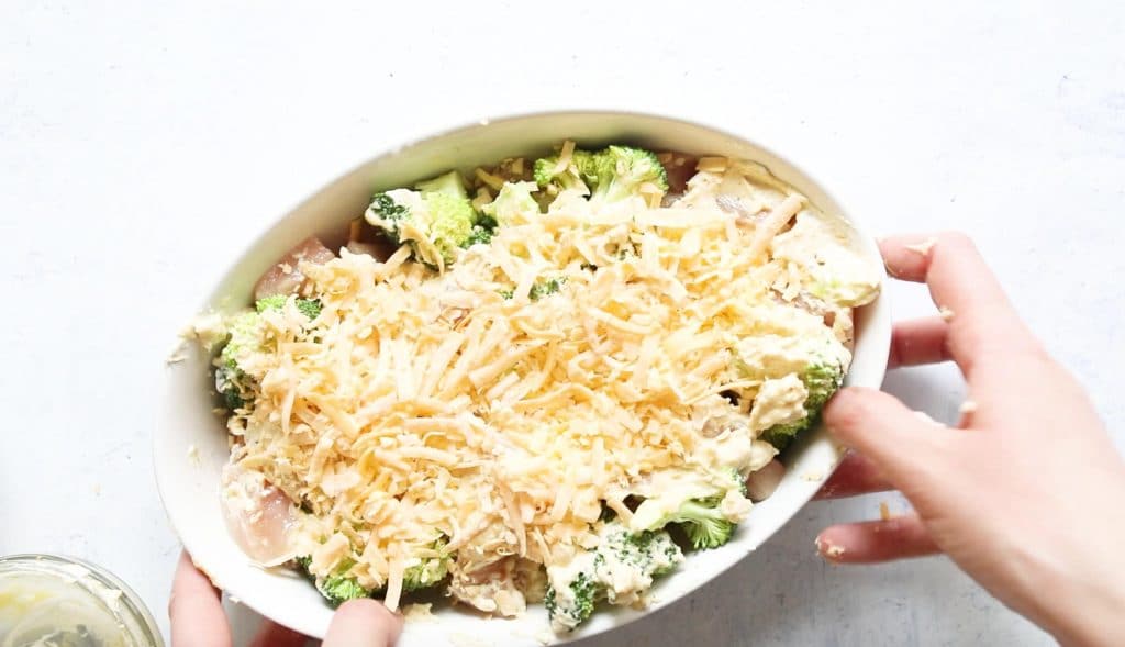 uncooked chicken and broccoli divan with cheese on top in a white oval baking dish