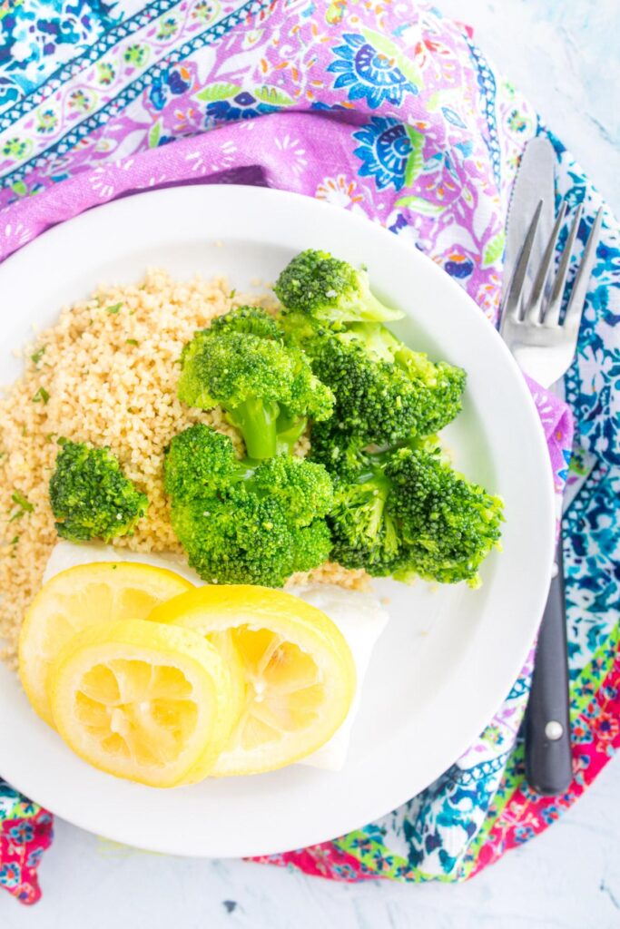 cod with lemon slices on top with couscous and broccoli on a white plate