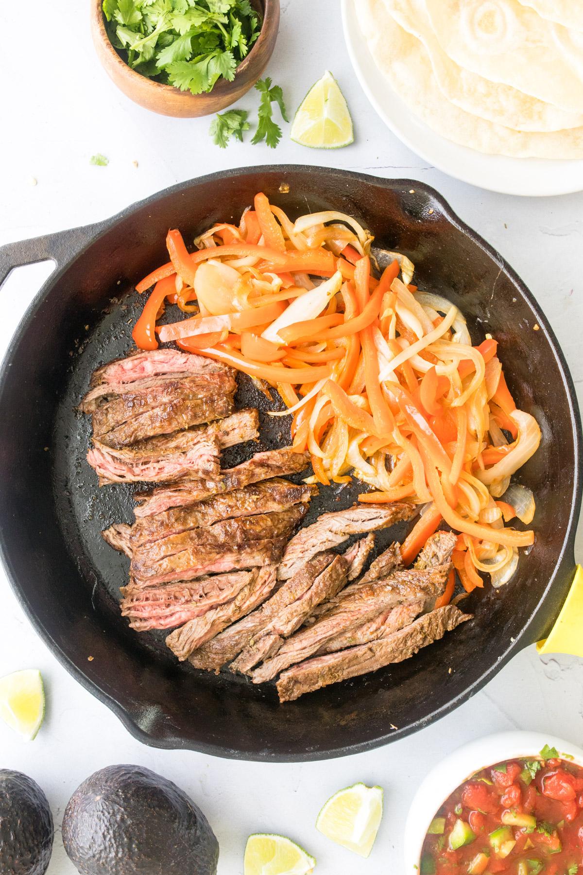 steak, peppers, and onions in a cast iron skillet surrounded by fajita ingredients