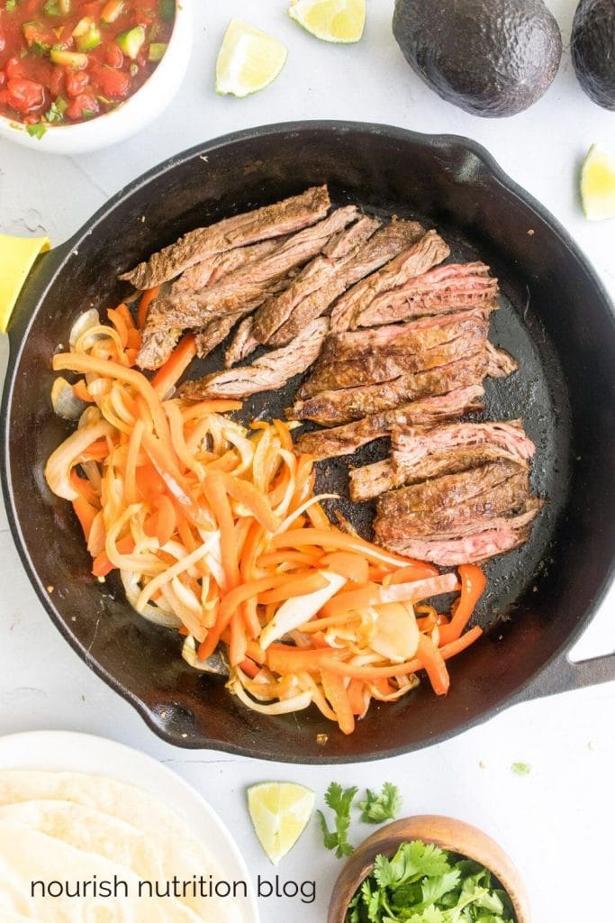 sliced steak and peppers/onions on a cast iron skillet surrounded by fajita toppings