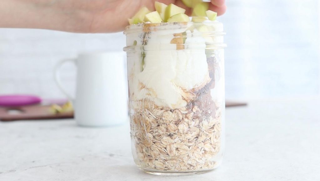 hand adding apples to a jar of overnight oats