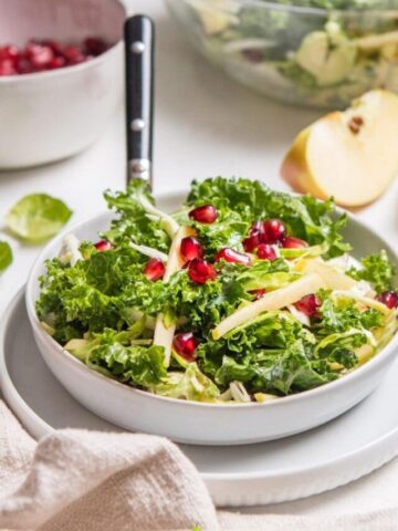 kale salad with pomegranates in white bow with ingredients behind it