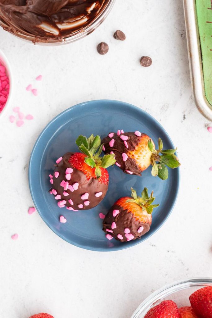 three chocolate covered strawberries with sprinkles on a blue plate with ingredients around it