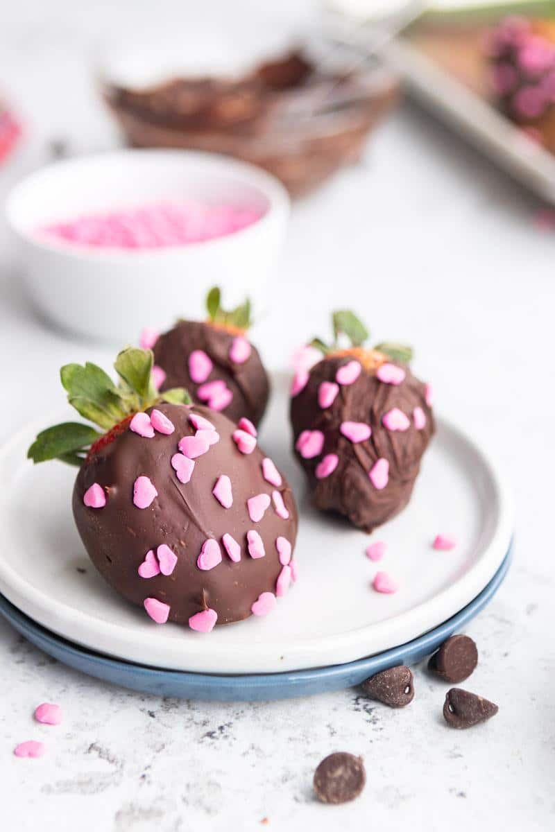 three chocolate covered strawberries with heart sprinkles on a plate with ingredients in background