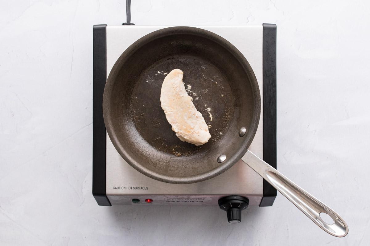 one chicken tender cooking in a black frying pan