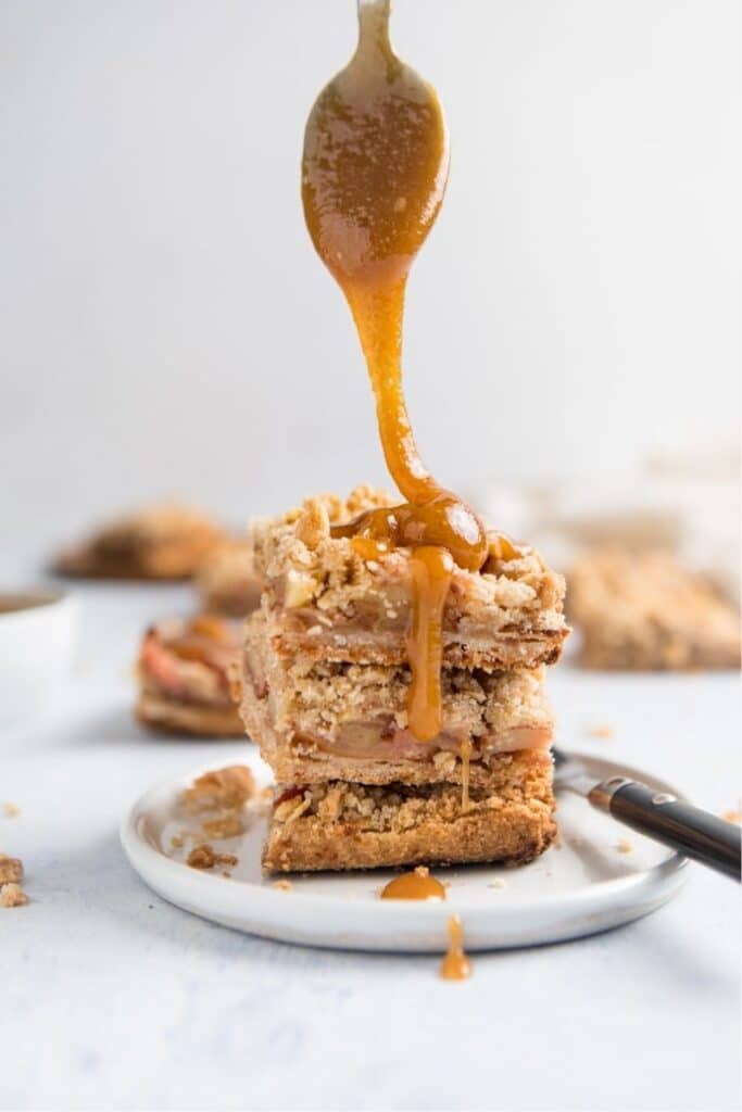 spoon drizzling caramel onto a stack of apple pie bars