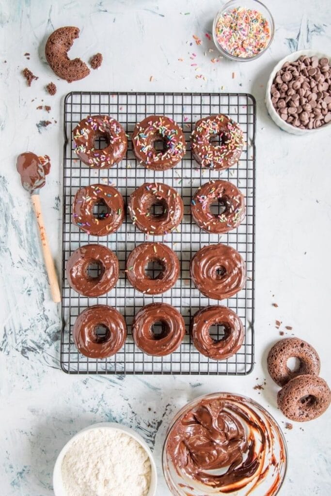 12 chocolate donuts, some with sprinkles on it with ingredients around it