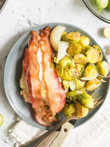 chicken with bacon on top next to brussels sprouts on a grey plate