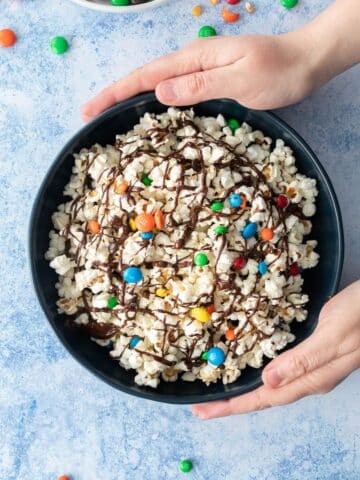 hands holding dark bowl of popcorn with m&ms and chocolate