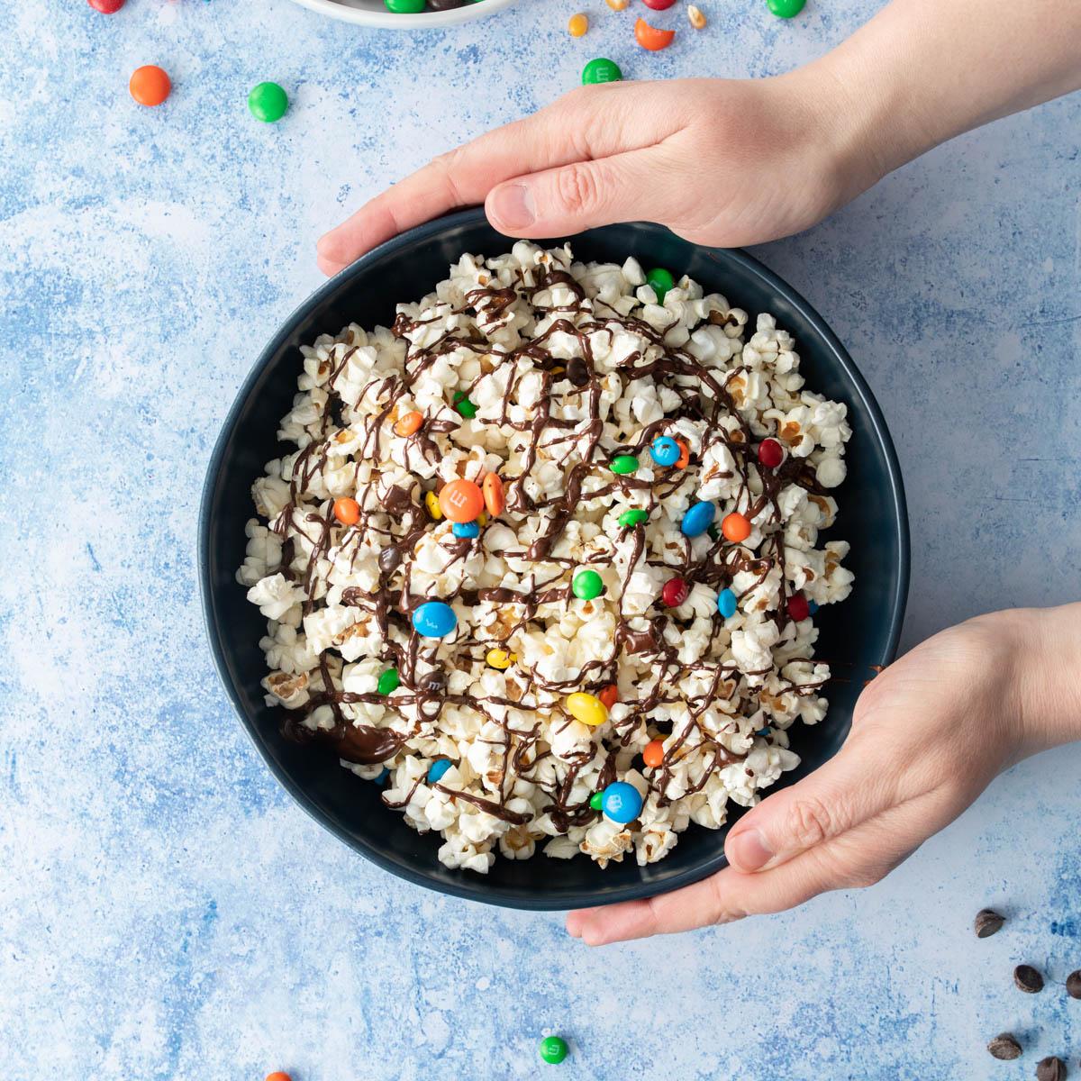 hands holding dark bowl of popcorn with m&ms and chocolate