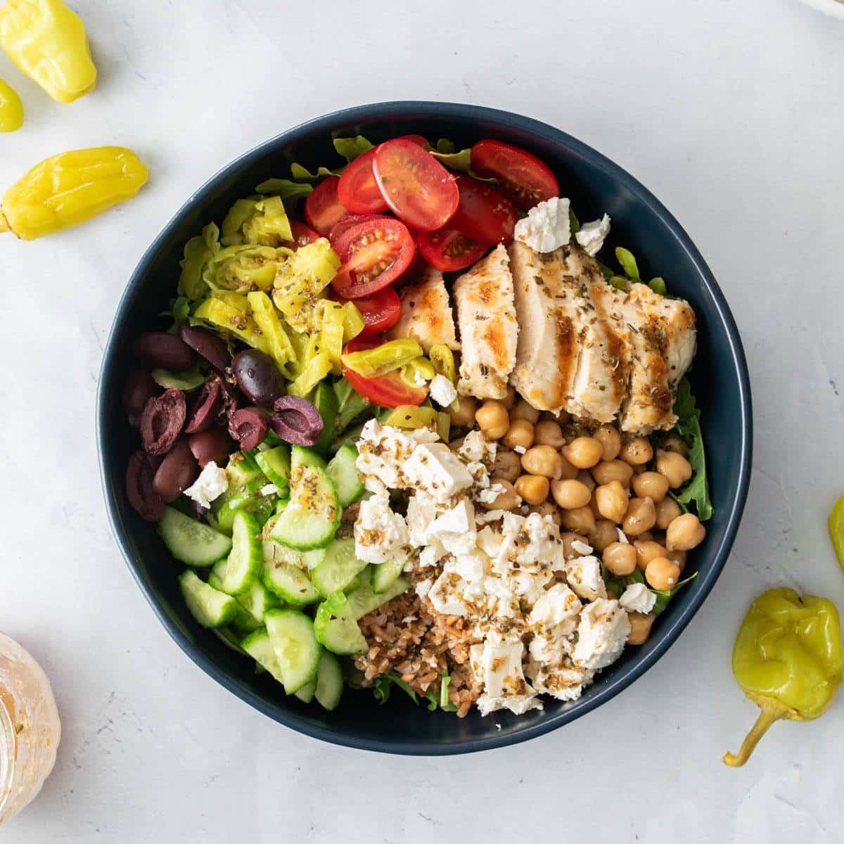 blue bowl full of vegetables, olives, grains, chickpeas, and chicken